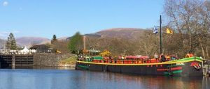 A colorful barge on a canal with a lock and mountains in the background under a blue sky.