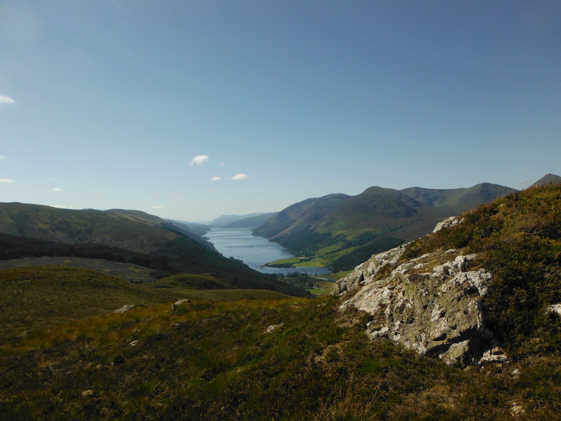 View of a long lake surrounded by green mountains and blue sky, with foreground of grass and rocks.
