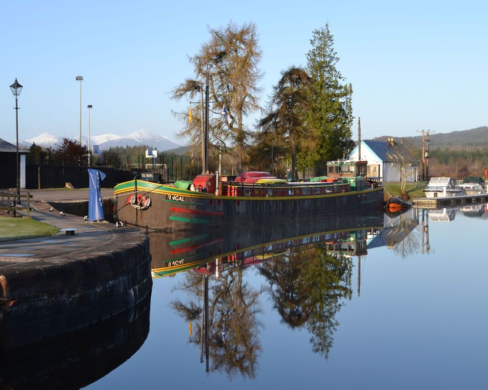 Barge on calm water, reflecting trees and sky. Mountains in the distance.