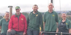 Five people stand outdoors: One wearing a red jacket and hat, the others in green shirts. Overcast day.