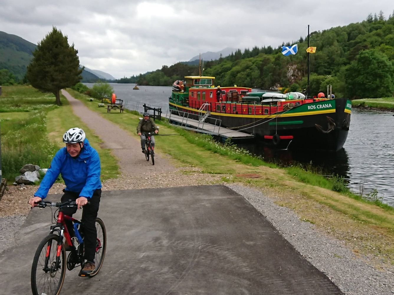 Cyclists on path beside canal, a colorful boat is docked. Green landscape, cloudy sky.