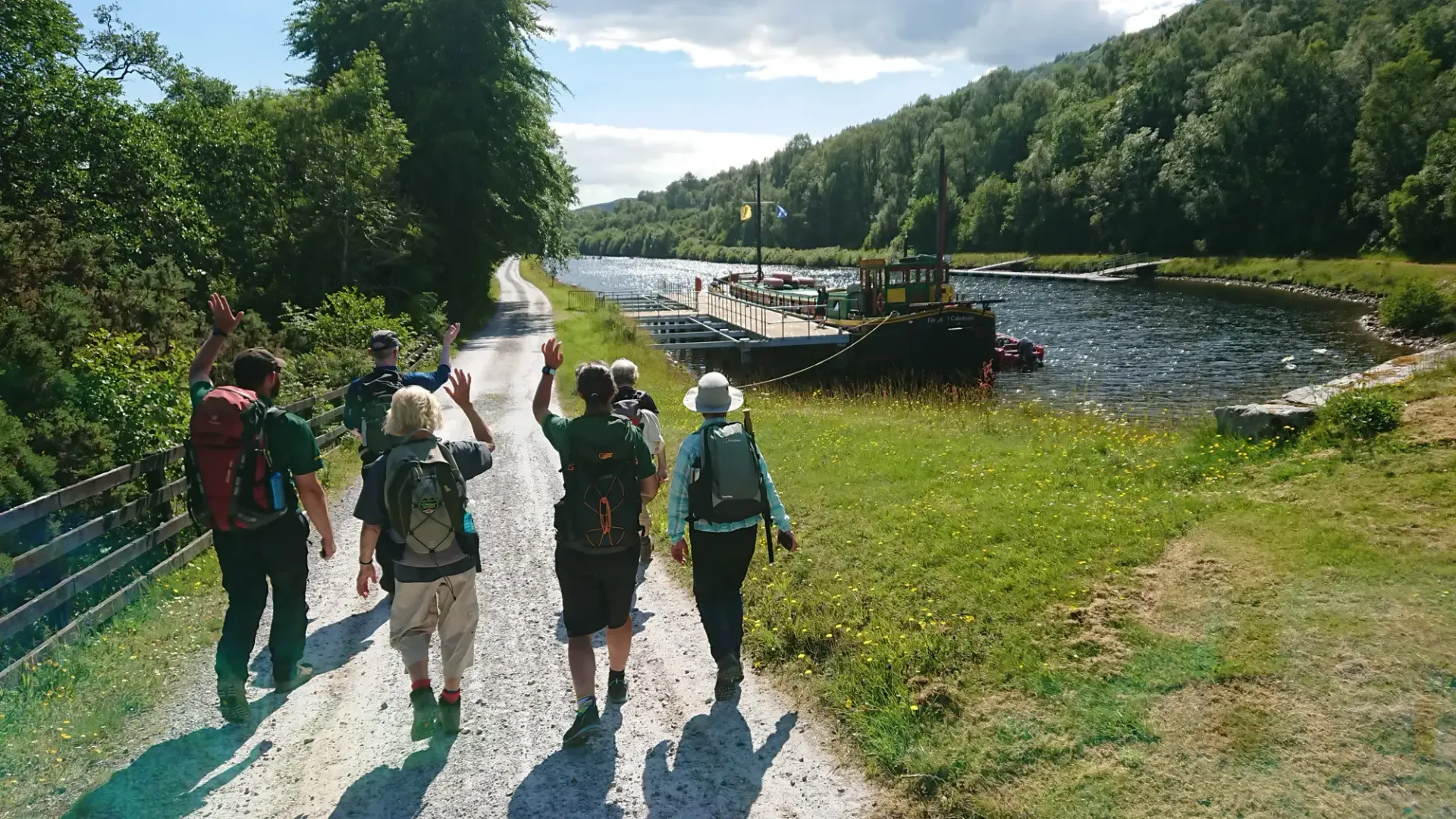 Group hiking along a gravel path next to a canal with a dock and boat, surrounded by trees.