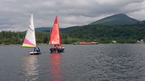 Two sailboats on a lake with a mountain and dark sky background. One has a red sail, one a white.