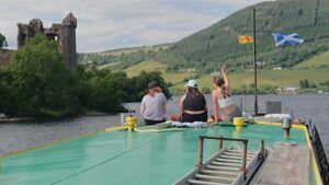 Three people on a boat with Urquhart Castle in the background, Scottish flags, and mountains.