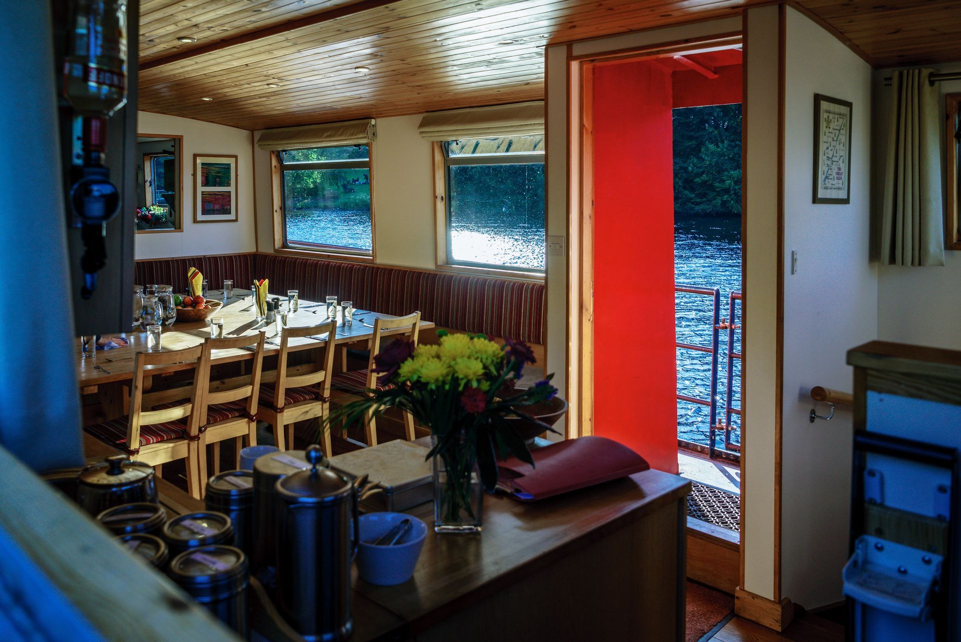 Interior of a boat with a dining table set for guests, a bright orange doorway, and windows overlooking water.