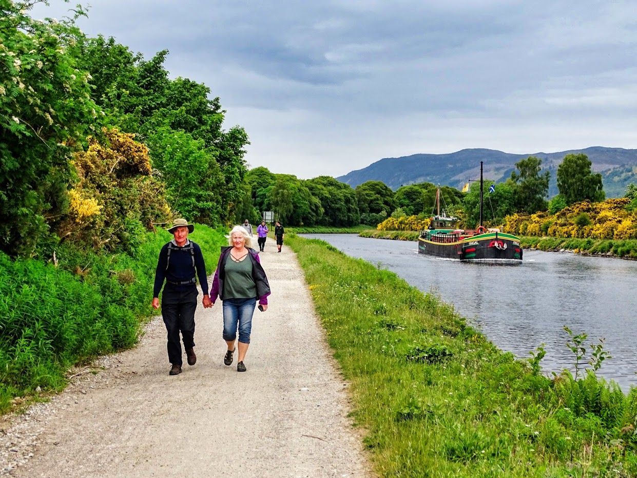 People walking on a path beside a canal, with a boat and greenery, under a cloudy sky.