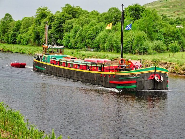 A long barge navigating a river, with green and red accents, flags, and lush green banks.