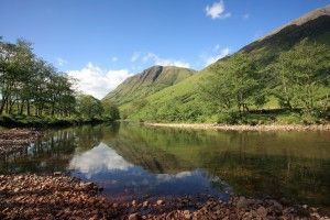 Calm river reflects mountains and trees under a clear blue sky.