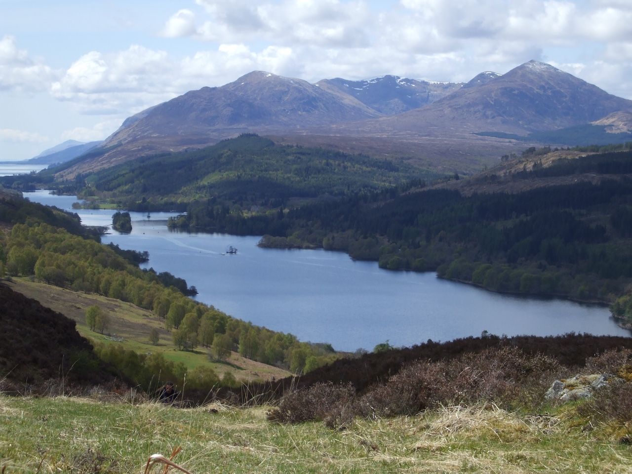 Loch in Scotland, surrounded by forested hills and mountains, under a partly cloudy sky.