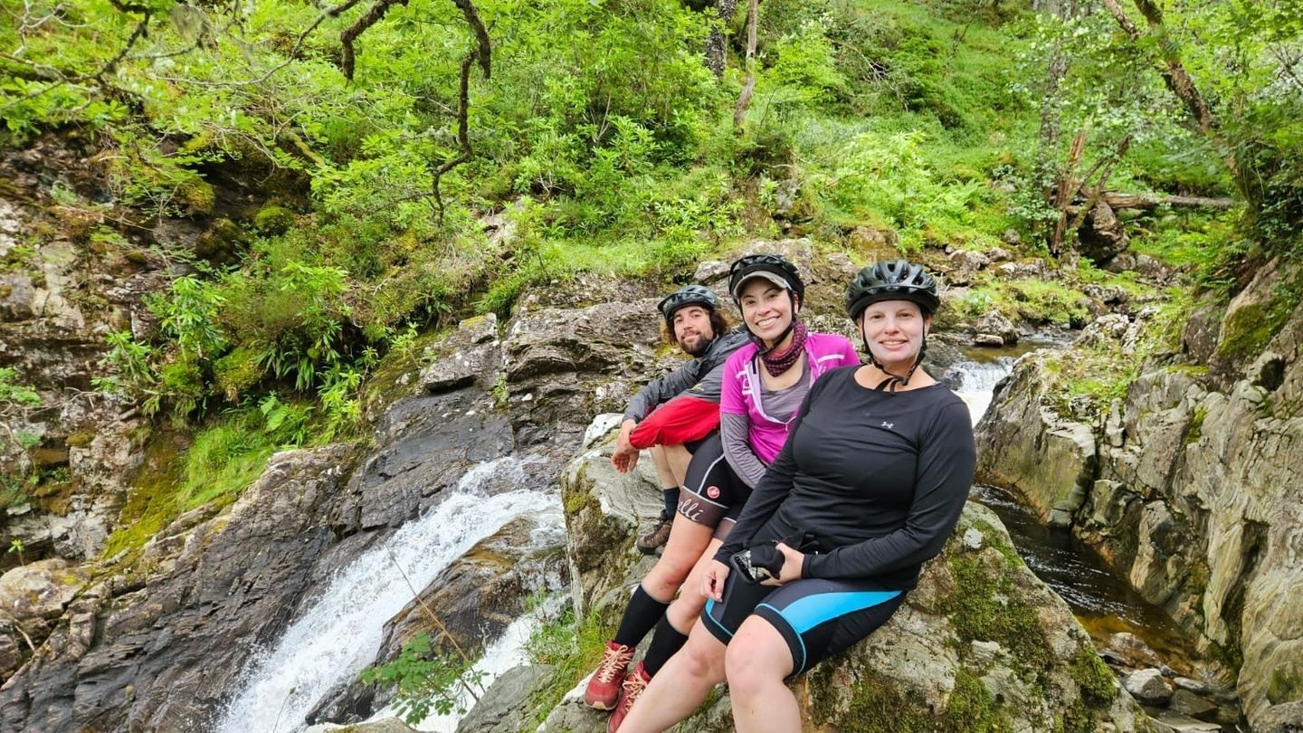 Three people in bike helmets resting by a waterfall in a lush, green environment.