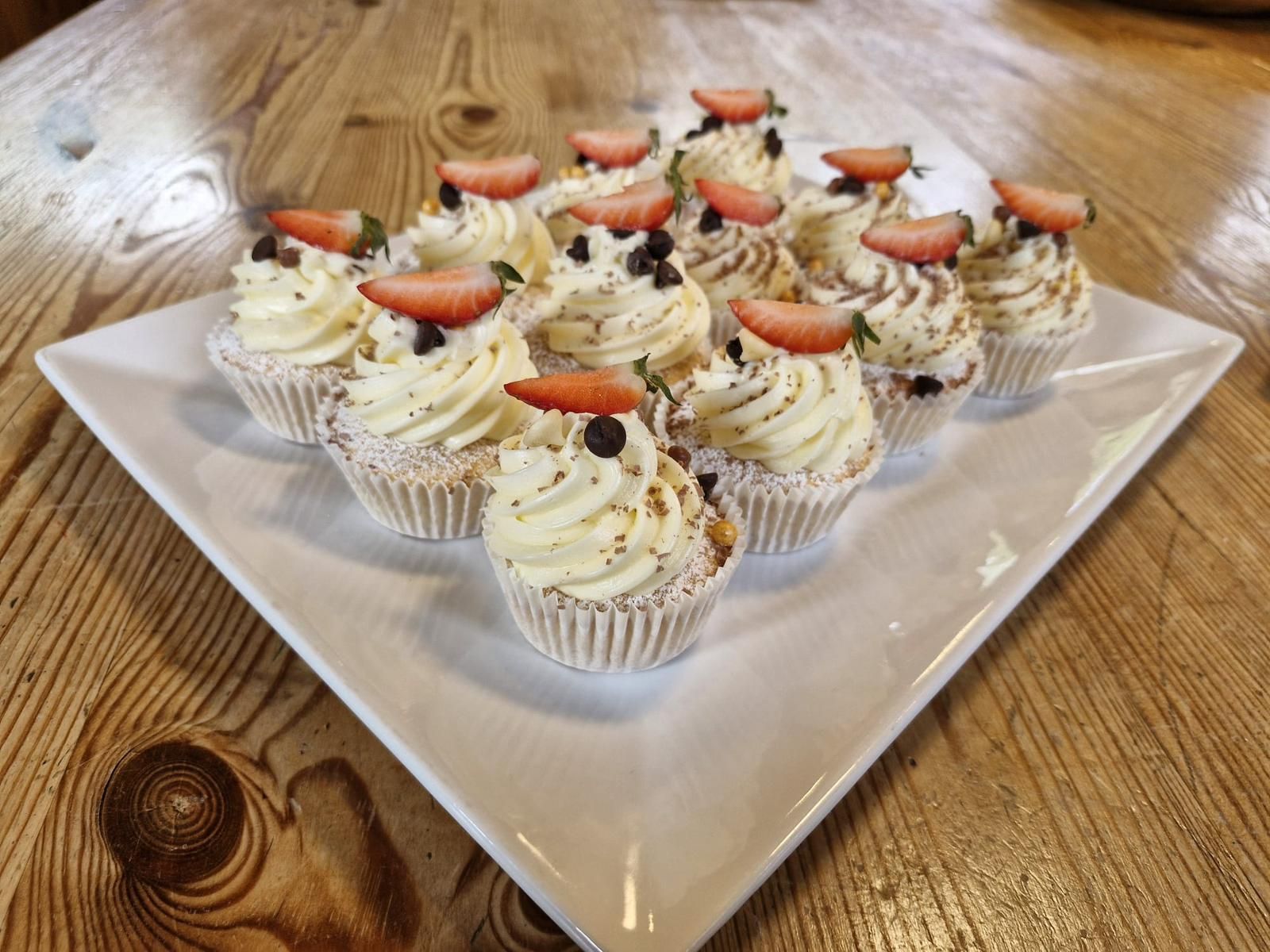 Cupcakes with cream frosting, chocolate chips, and strawberry slices, on a white square platter, set on a wooden table.