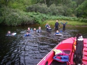 People in wetsuits swimming in a dark lake near a red boat. Dog in the boat wearing a life vest.