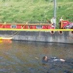 Person swimming in water next to a red and yellow boat, green hillside in the background.