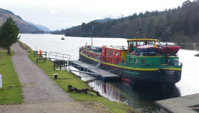 Colorful boat docked on a lake with a path, trees, and mountains in the background.