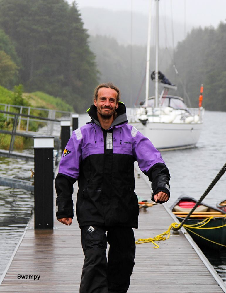 Man in purple/black jacket walks on a wooden dock with a sailboat in the background.