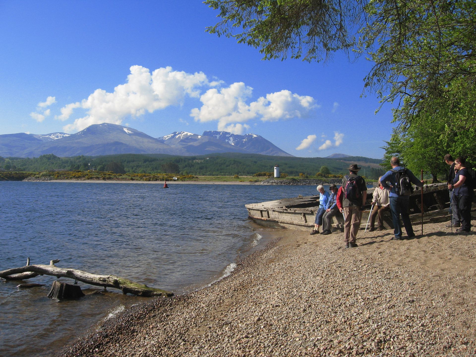 Lake shore with people, mountains in the distance, blue sky with clouds.