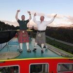 Two men in kilts with arms raised atop a red boat, against a sunset.