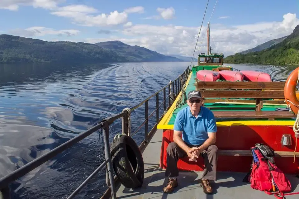 Man seated on ferry deck, looking at the water. Loch Ness, Scotland with mountains in background. Blue sky and sunny.