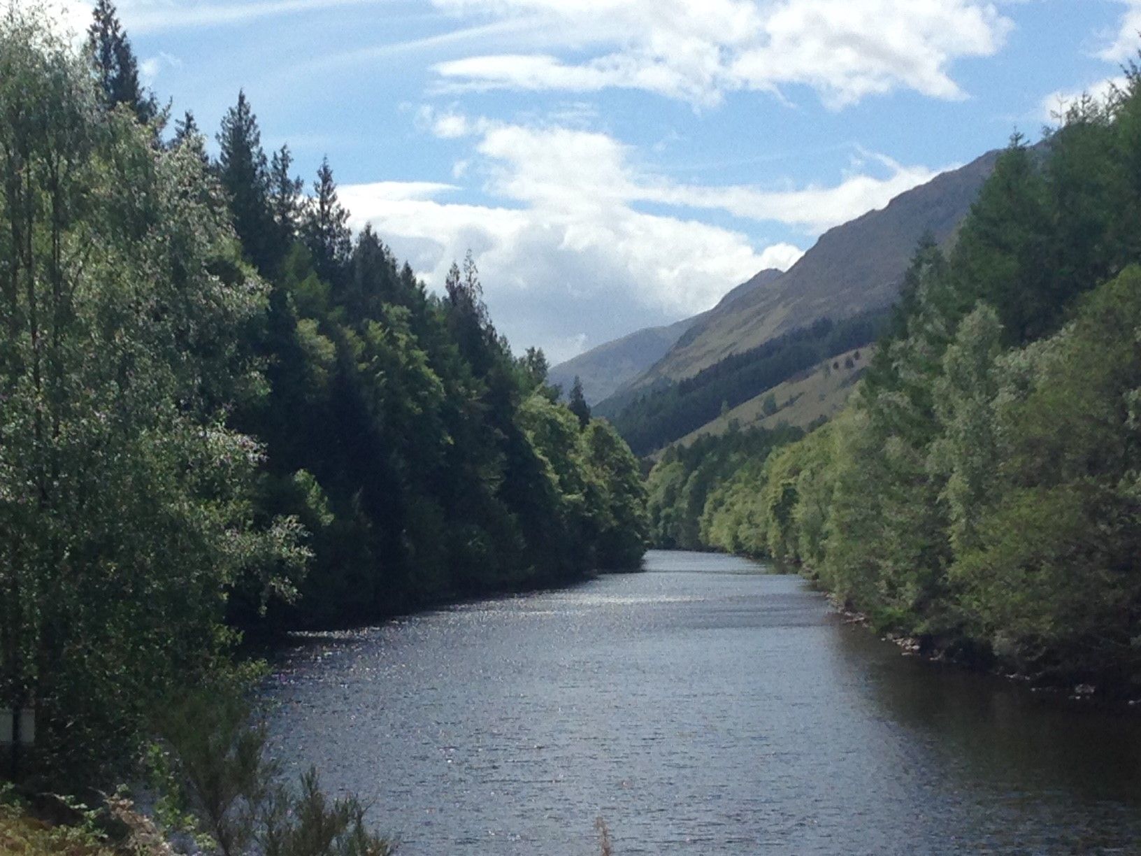 River winding through a valley, flanked by trees and mountains under a partly cloudy sky.