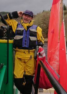 Man in yellow sailing gear, on a boat with red sail, adjusting hat, smiling.