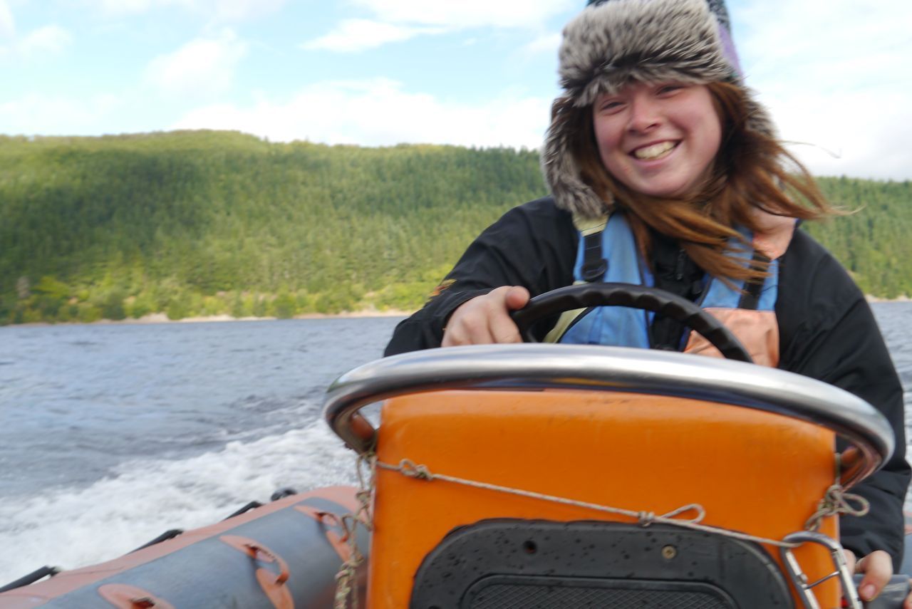 Person smiling, driving a boat on water. Wearing furry hat and life vest. Forest in background.
