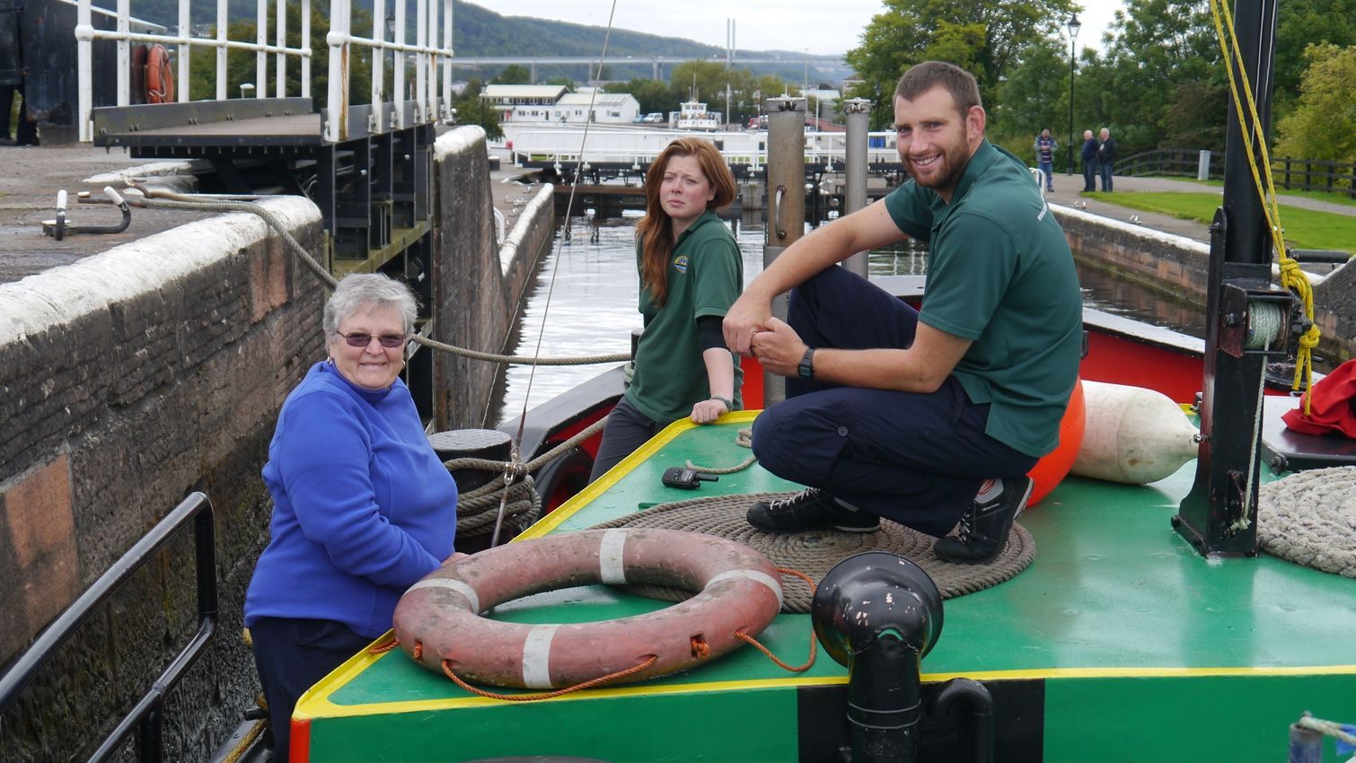Three people on a green boat in a canal lock. One woman stands, two others sit, looking at the camera. A bridge is in the background.