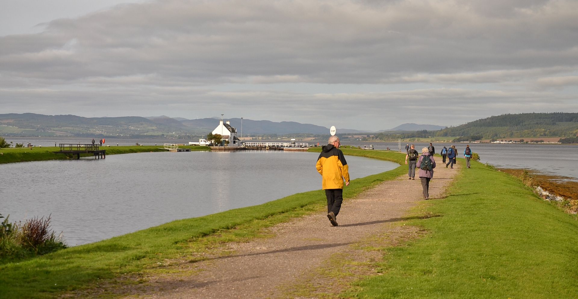 People walking on a path along a body of water toward a building on the water, under a cloudy sky.