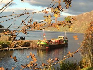 Boat docked on a lake, framed by branches with autumn leaves. Mountain in the background.