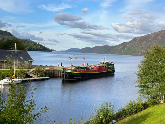 Loch Ness cruise boat docked at pier, Scottish Highlands. Water, hills, blue sky with clouds.