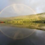 Full rainbow arching over a calm lake, reflected in the water, against a green hill and cloudy sky.
