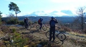 Cyclists pause on a trail, looking at snowy mountains under a blue sky.