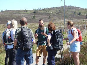 Group of hikers on a path, listening to a man with a beard holding an object; sunny landscape background.