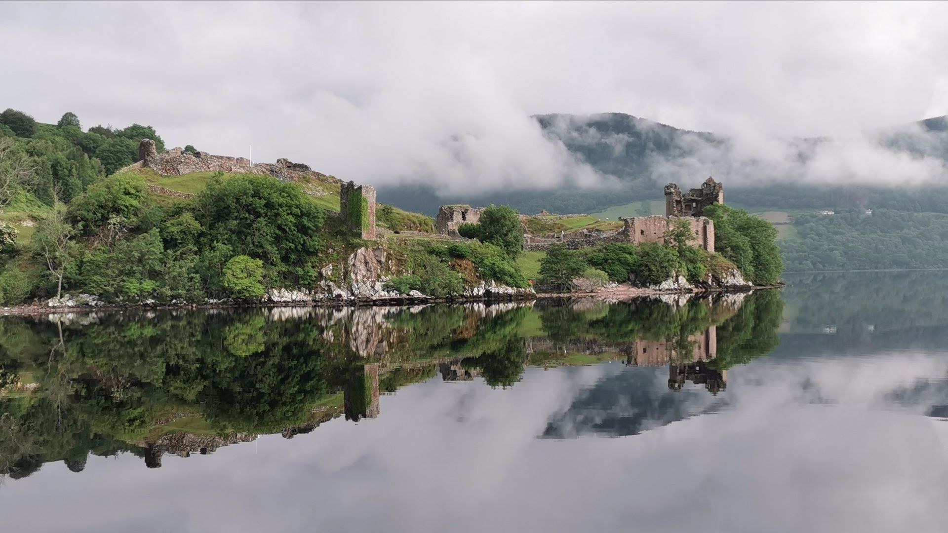 Ruins of Urquhart Castle on Loch Ness with water reflection, under a cloudy sky.