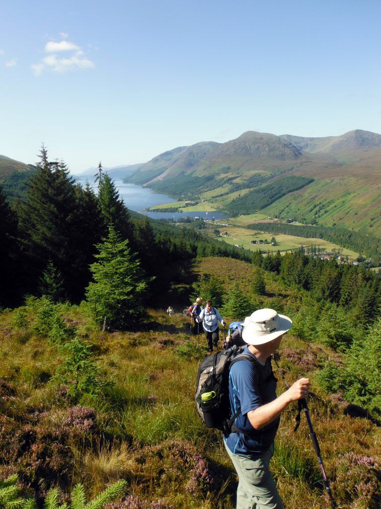 Hikers with backpacks on a hillside path, overlooking a valley with a lake and mountains. Sunny day.
