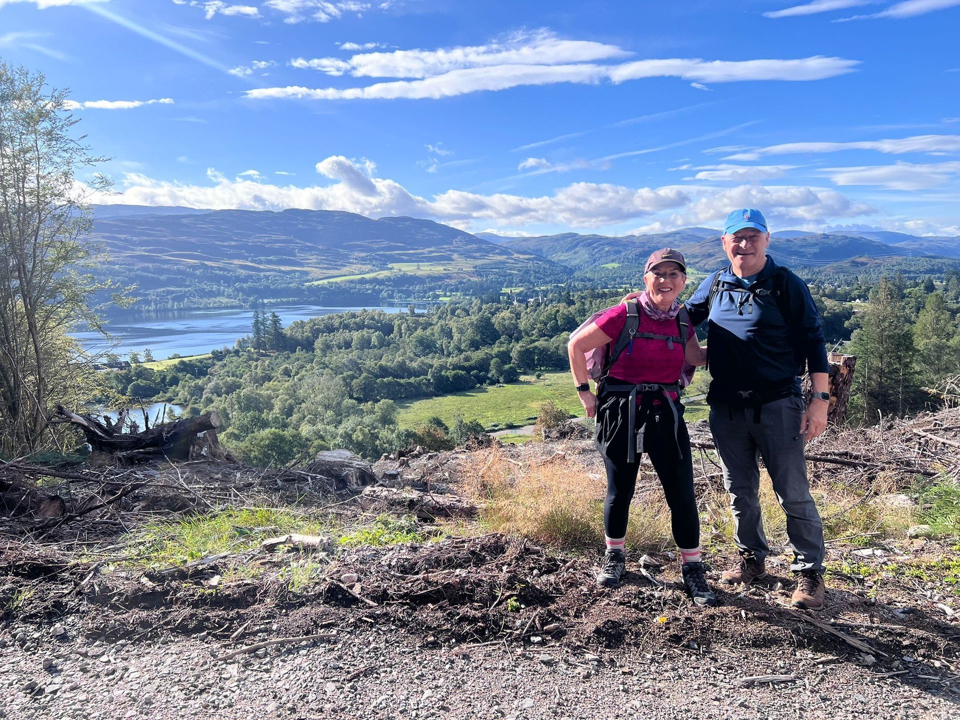 Two hikers pose overlooking a valley and lake under a blue sky with clouds.