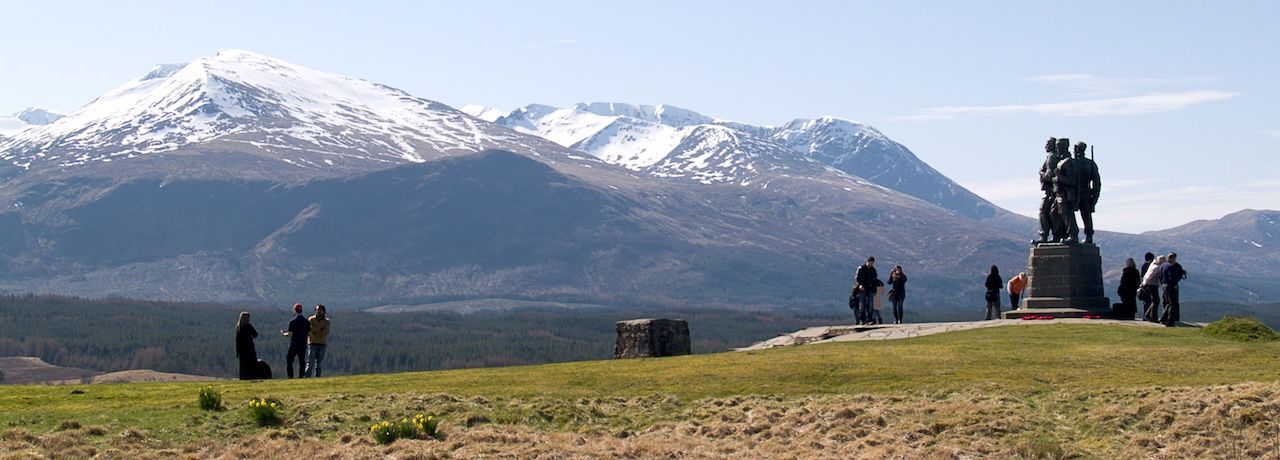 Monument on a grassy hill with snowy mountains in the background. People stand near monument and in the distance.