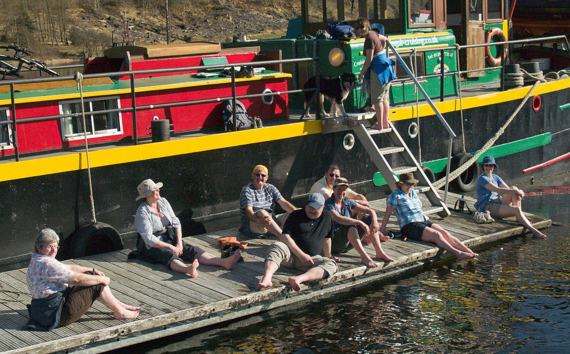 People sunbathing on a dock beside a colorful canal boat with a ladder.