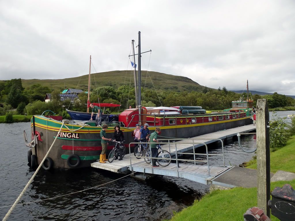 Barge docked at a pier, people with bicycles, water and a hill in the background.