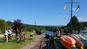Cyclists by a canal with colorful kayaks. Blue sky, green trees, and distant hills.