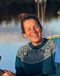 Woman smiling outdoors, wearing teal patterned sweater, water in background.
