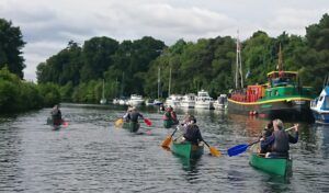 People paddling canoes on a river with boats docked along the shore. Green canoes, trees, cloudy sky.