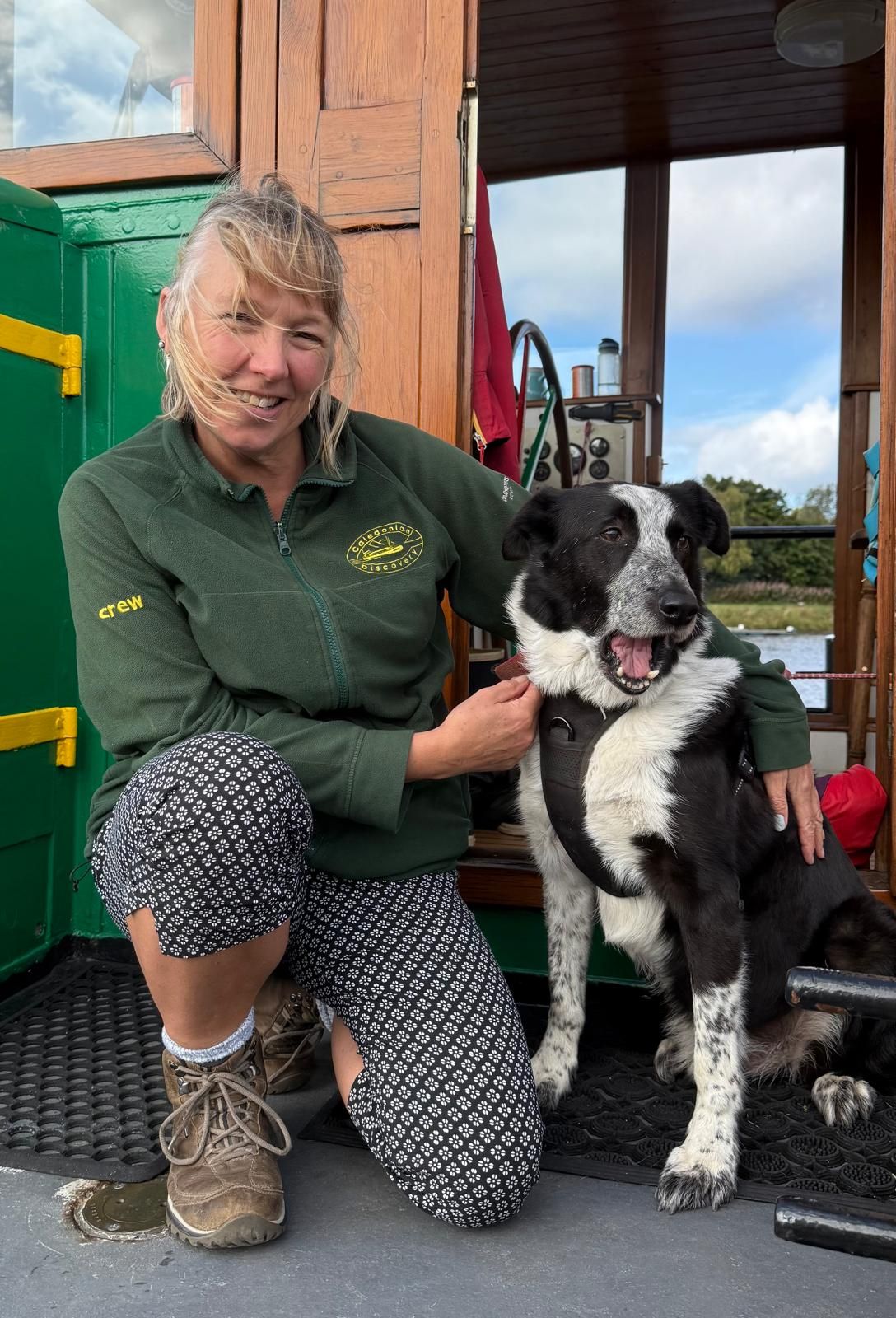 Woman kneels beside a black and white dog on a boat. The woman wears green shirt and patterned pants, smiling, petting the dog.