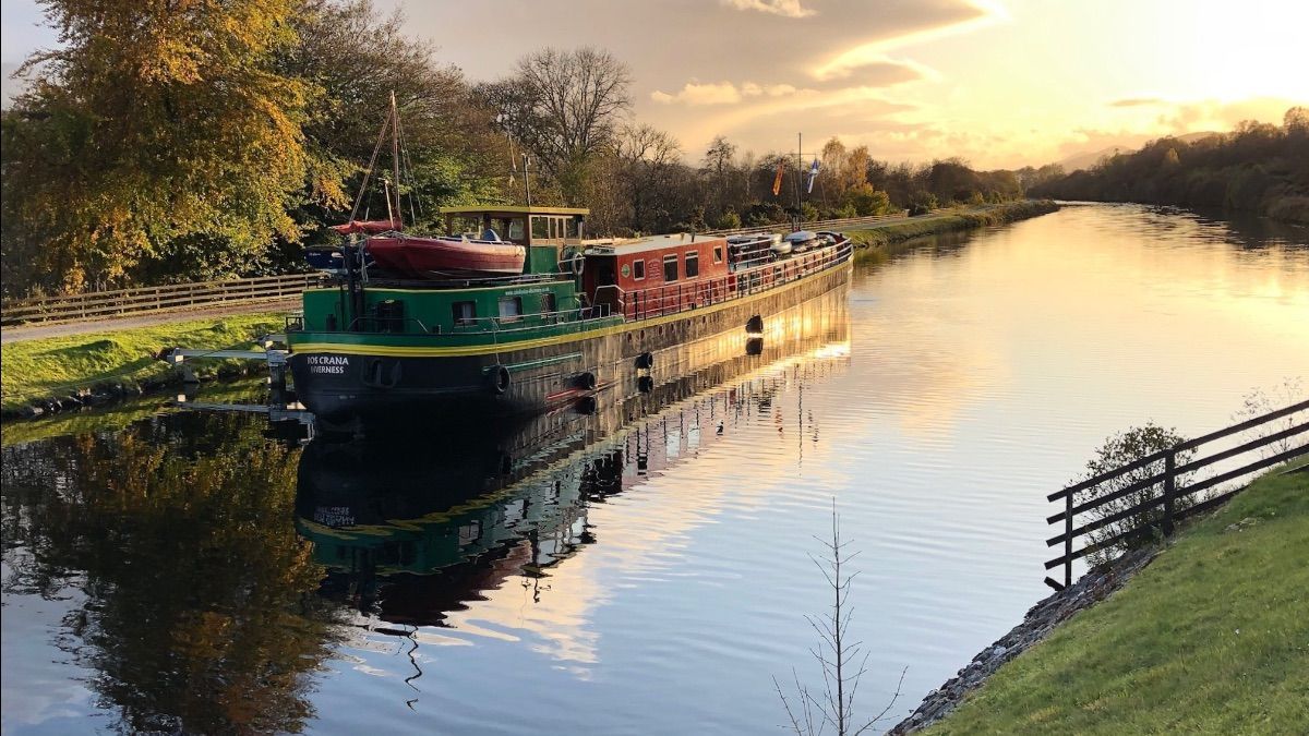 Barge on a calm river reflects the sunset. Green and red hull, trees line the banks.