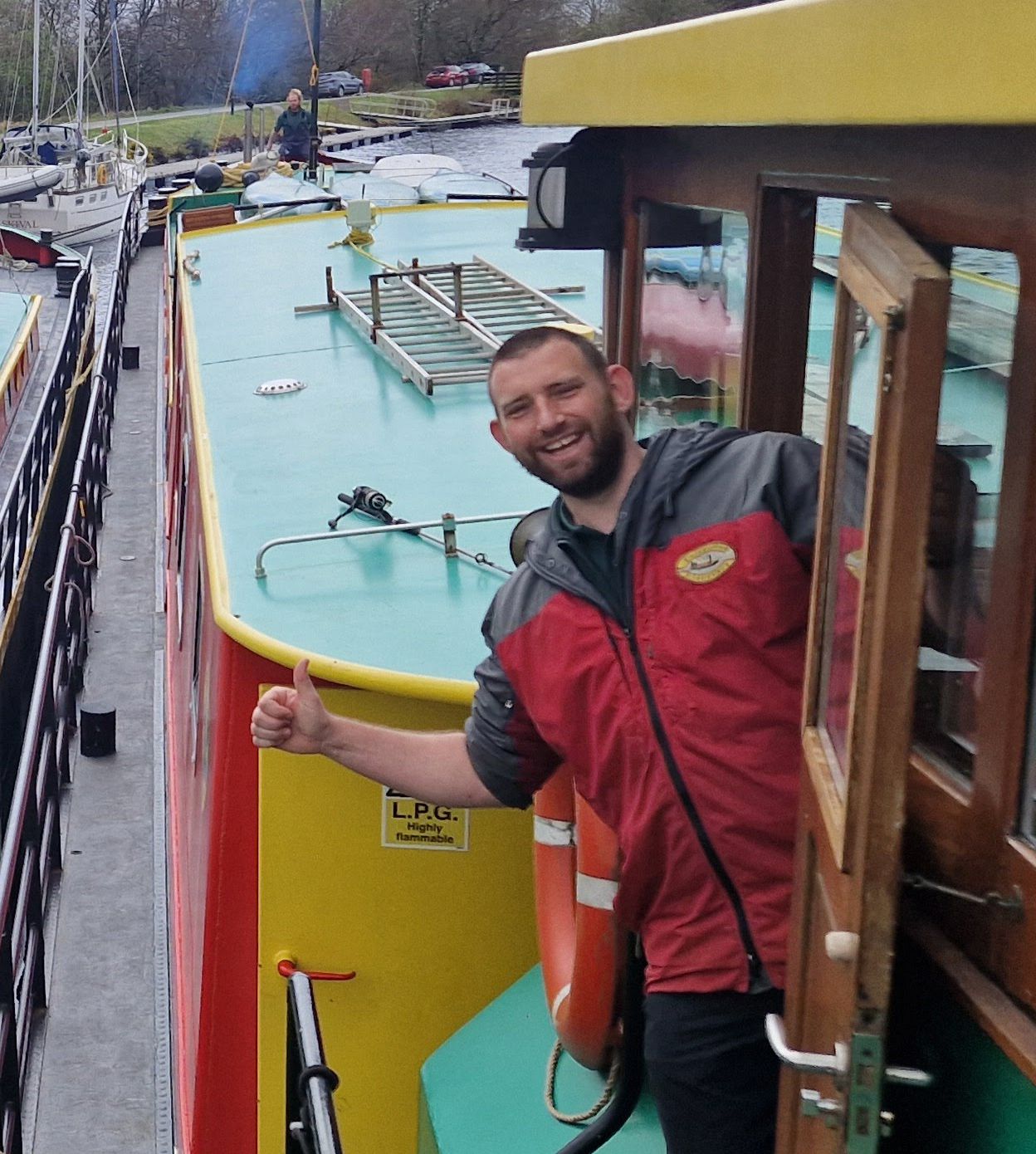 Man in red jacket gives a thumbs-up from a colorful canal boat door.