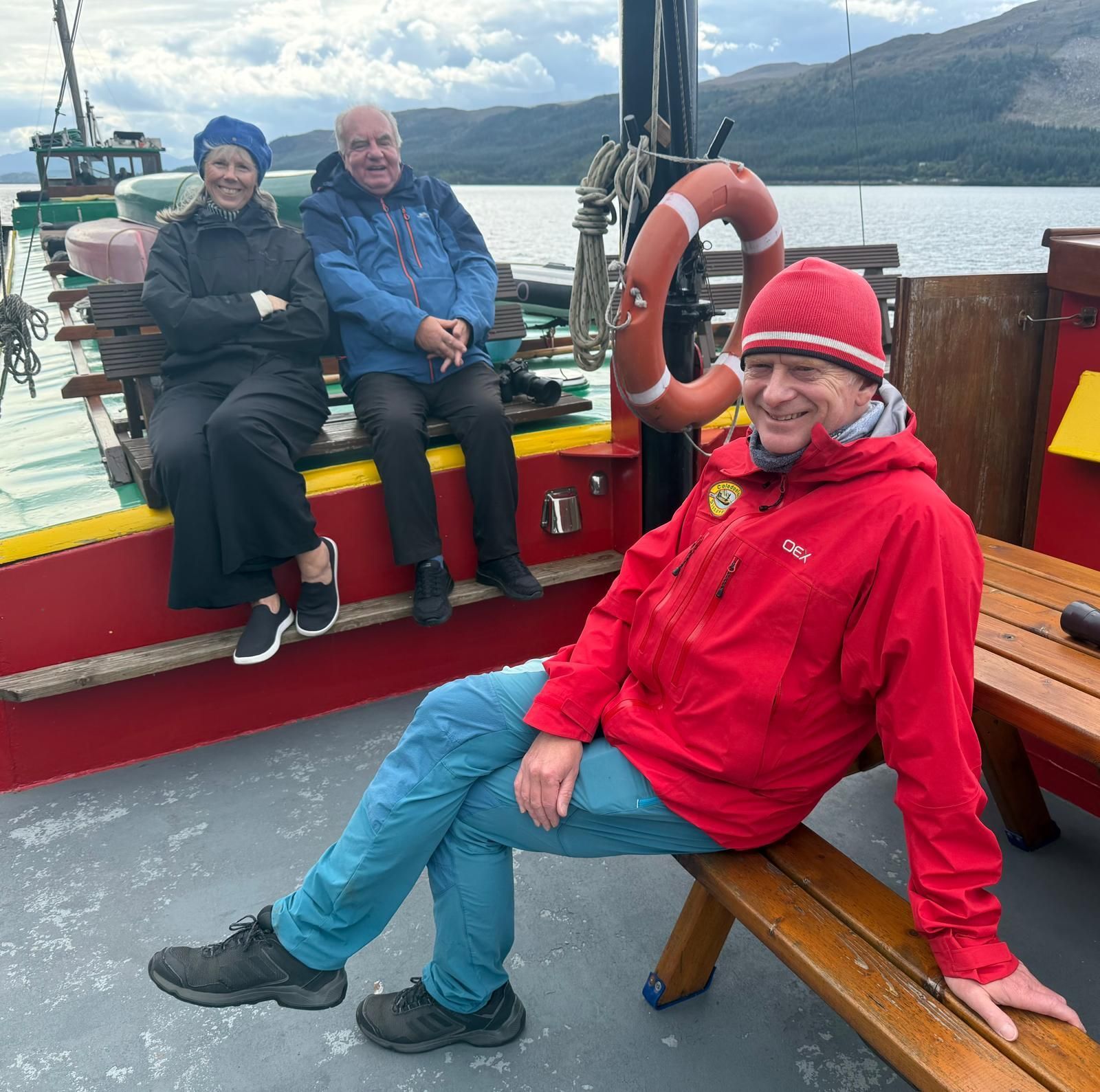 Three people on a boat deck, smiling at the camera. One sits with crossed arms; another, in red jacket, leans. Water and mountains in background.