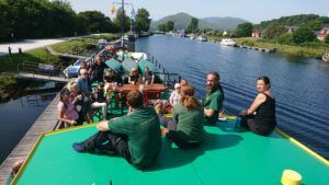 People on a green-decked boat on a canal, some seated around a table, enjoying the day. Trees and buildings line the waterway.