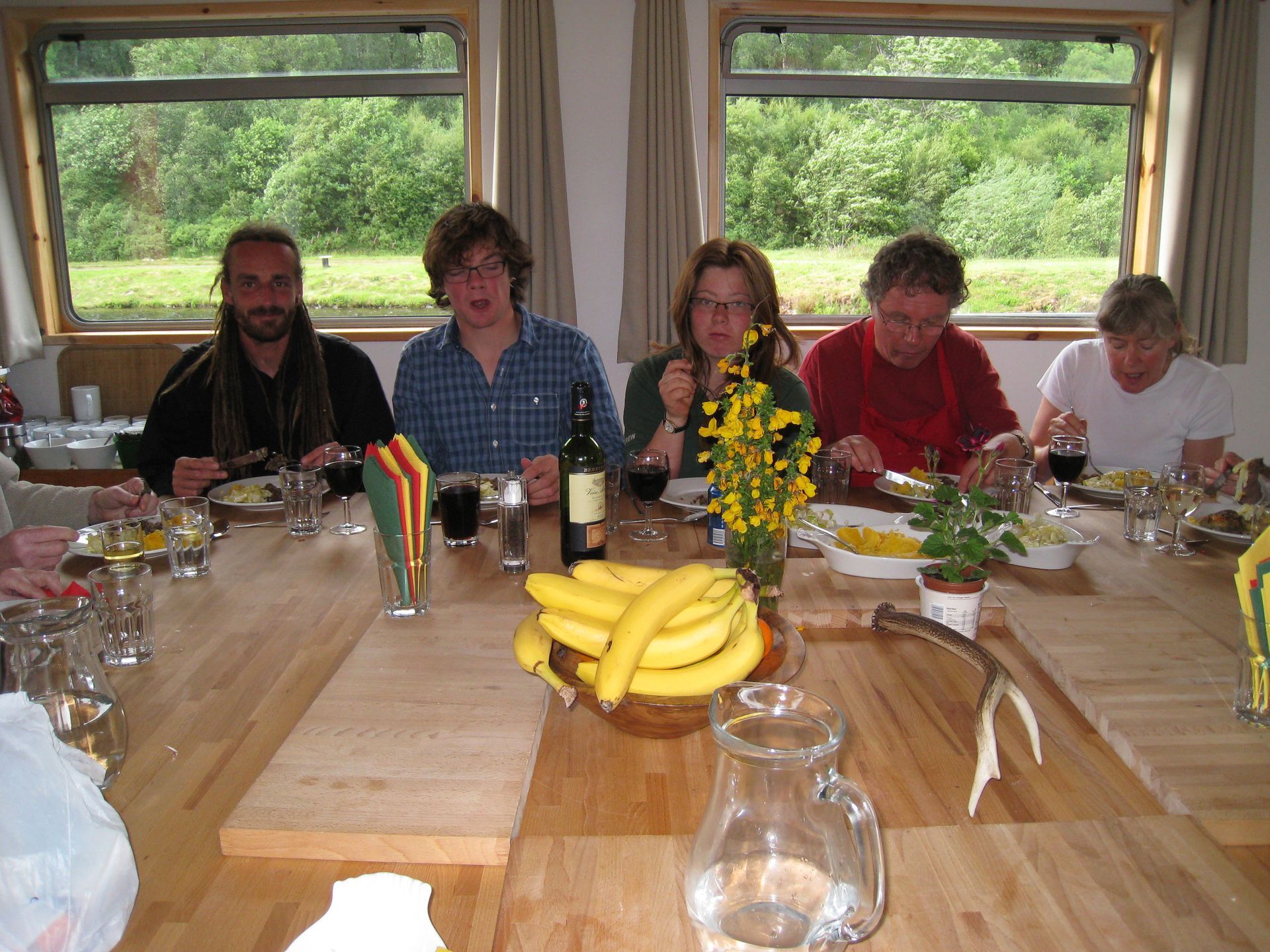 People eating at a long table inside a boat, surrounded by windows. Bananas and a flower bouquet are on the table.