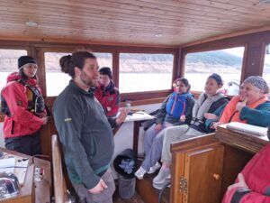 Group on a boat listening to a man speaking, likely giving a tour. Indoors, winter wear.