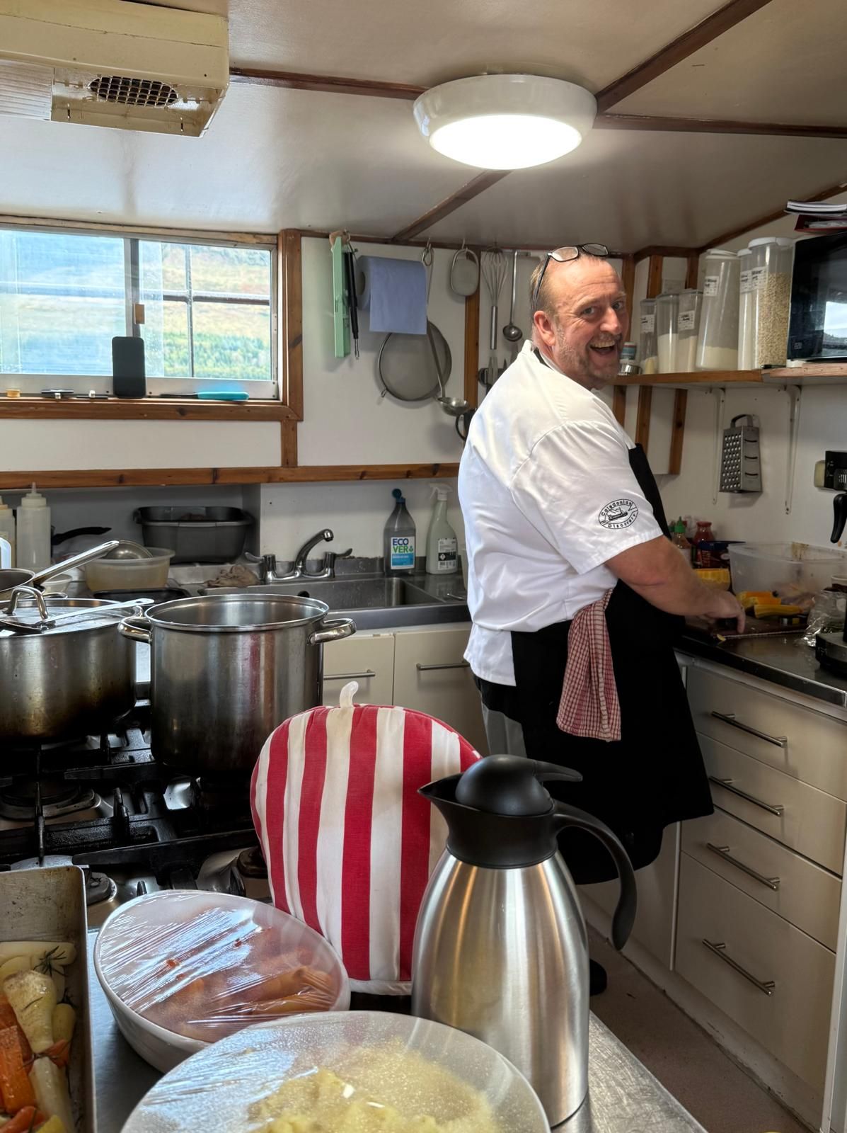 Chef in a kitchen, wearing a white shirt and black apron, smiling while working at a countertop.