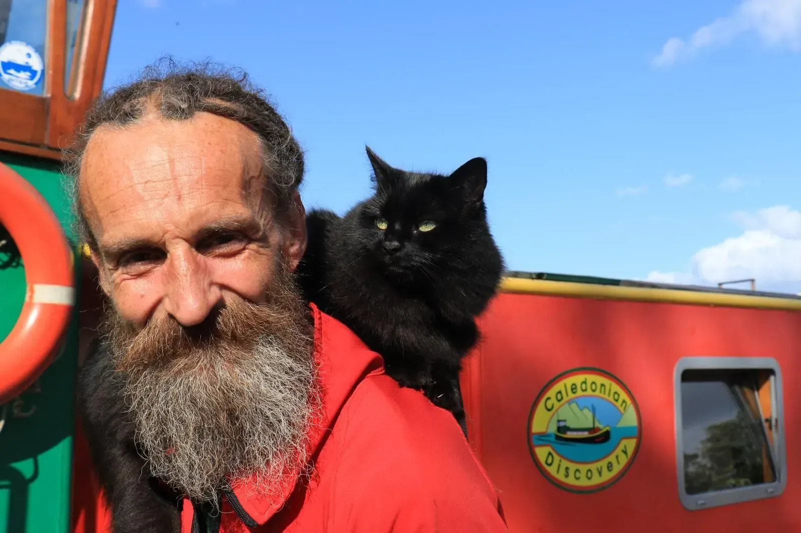 Man with a long beard and a black cat perched on his shoulder, against a red boat.